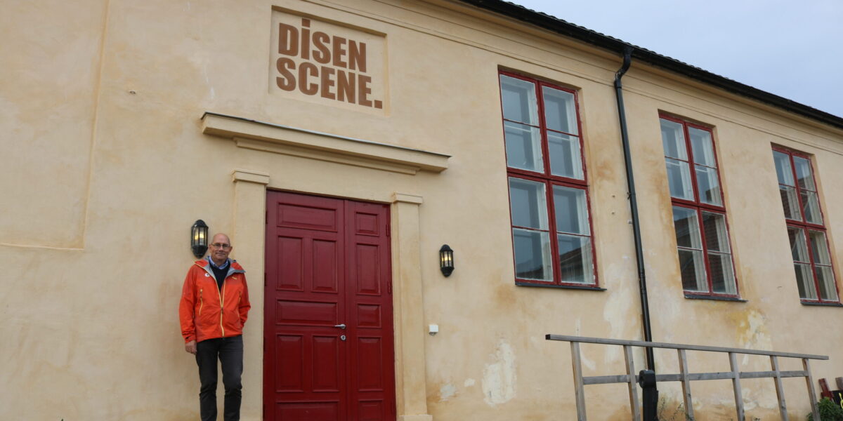 Bildet viser gult bygg. Rød tekst over døren er: Disen Scene. Styreleder står ved siden av døren.