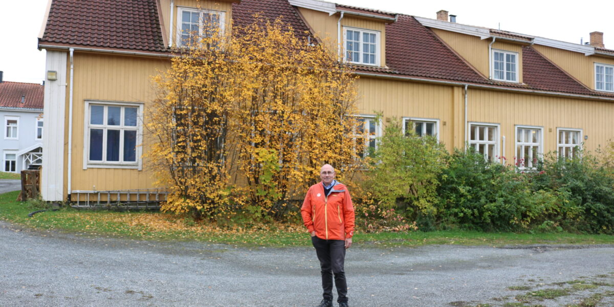 Bildet viser et gult, langt hus. Mann i front er styreleder med oransj jakke.