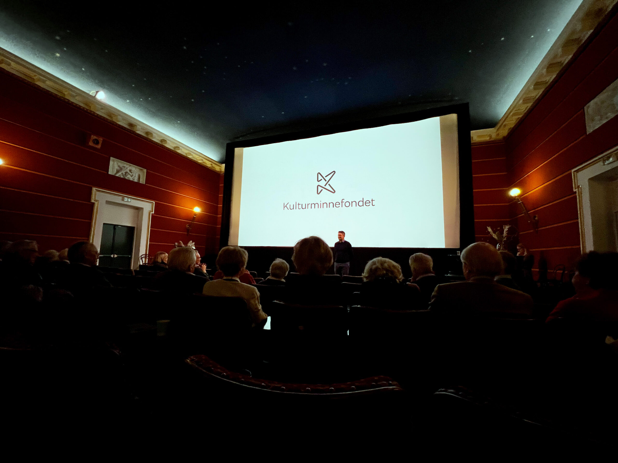 Bilde av unnsiden av kinoen, med publiku. På skjermer er Kulturminnefondets logo og teksten "Kulturminnefondet": 